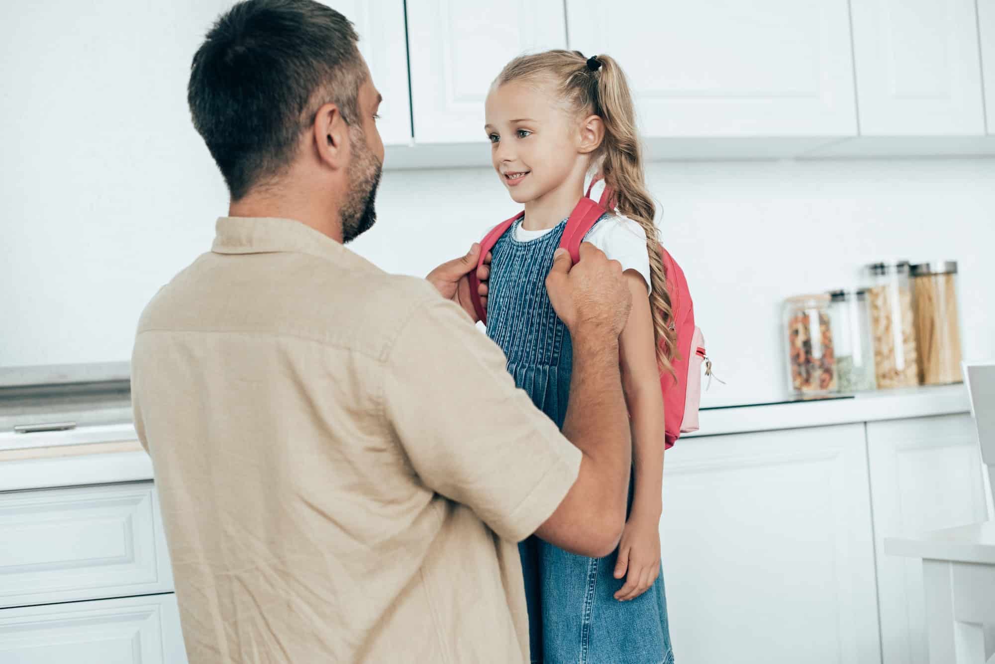 portrait of smiling father and little daughter with backpack at home, back to school concept